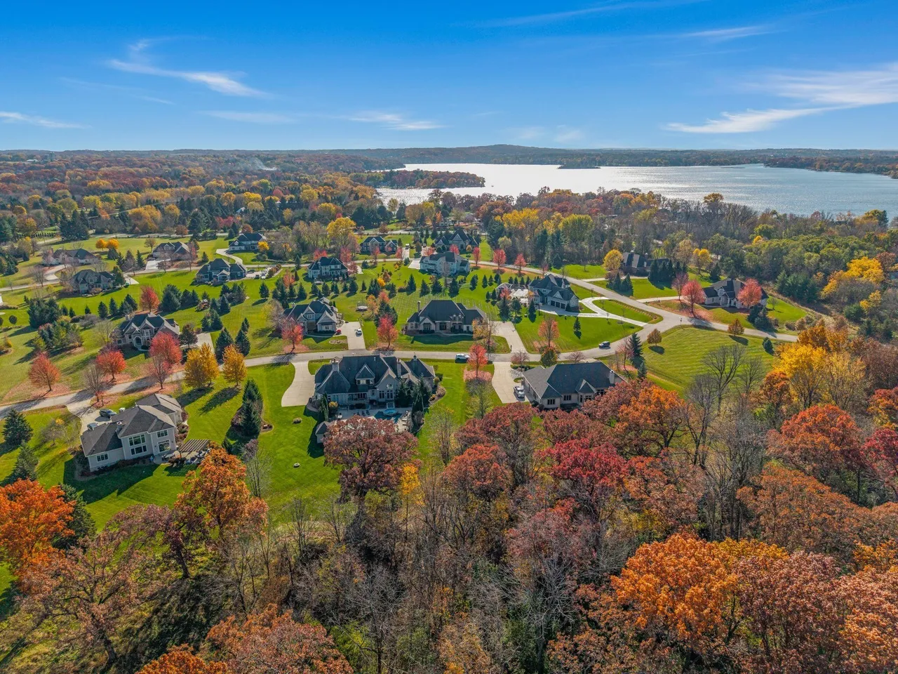 Aerial view of a scenic residential area, showcasing expansive homes surrounded by lush green lawns and mature trees in autumn colors. A serene lake is visible in the background, adding to the property's value and appeal. The community exudes a sense of luxury, privacy, and natural beauty, making it an attractive location for potential buyers.