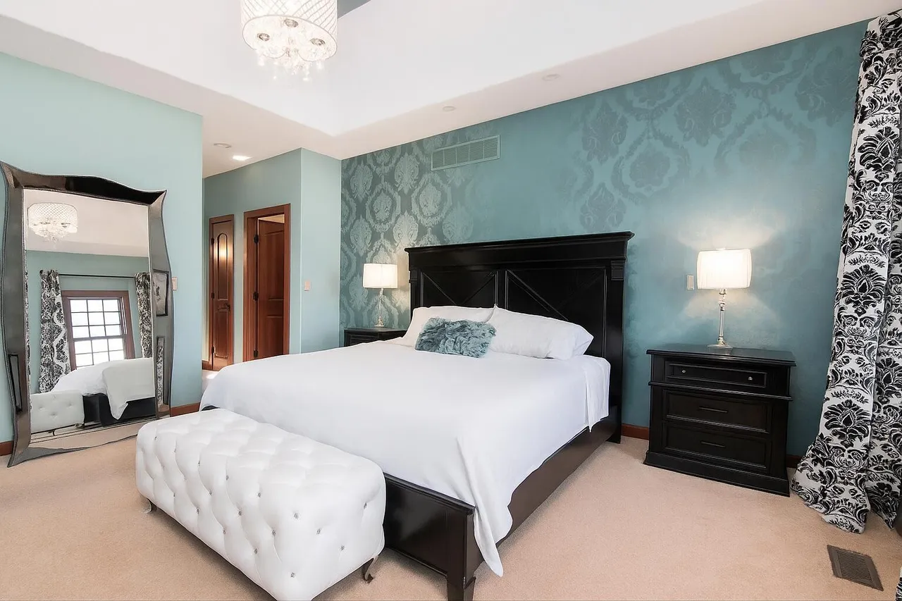 This is a primary bedroom featuring a king-sized bed with a black headboard, crisp white linens, and a decorative throw pillow. The walls are painted in a calming blue with a subtle damask pattern on the accent wall. A large mirror and tufted bench add to the room's luxurious feel.