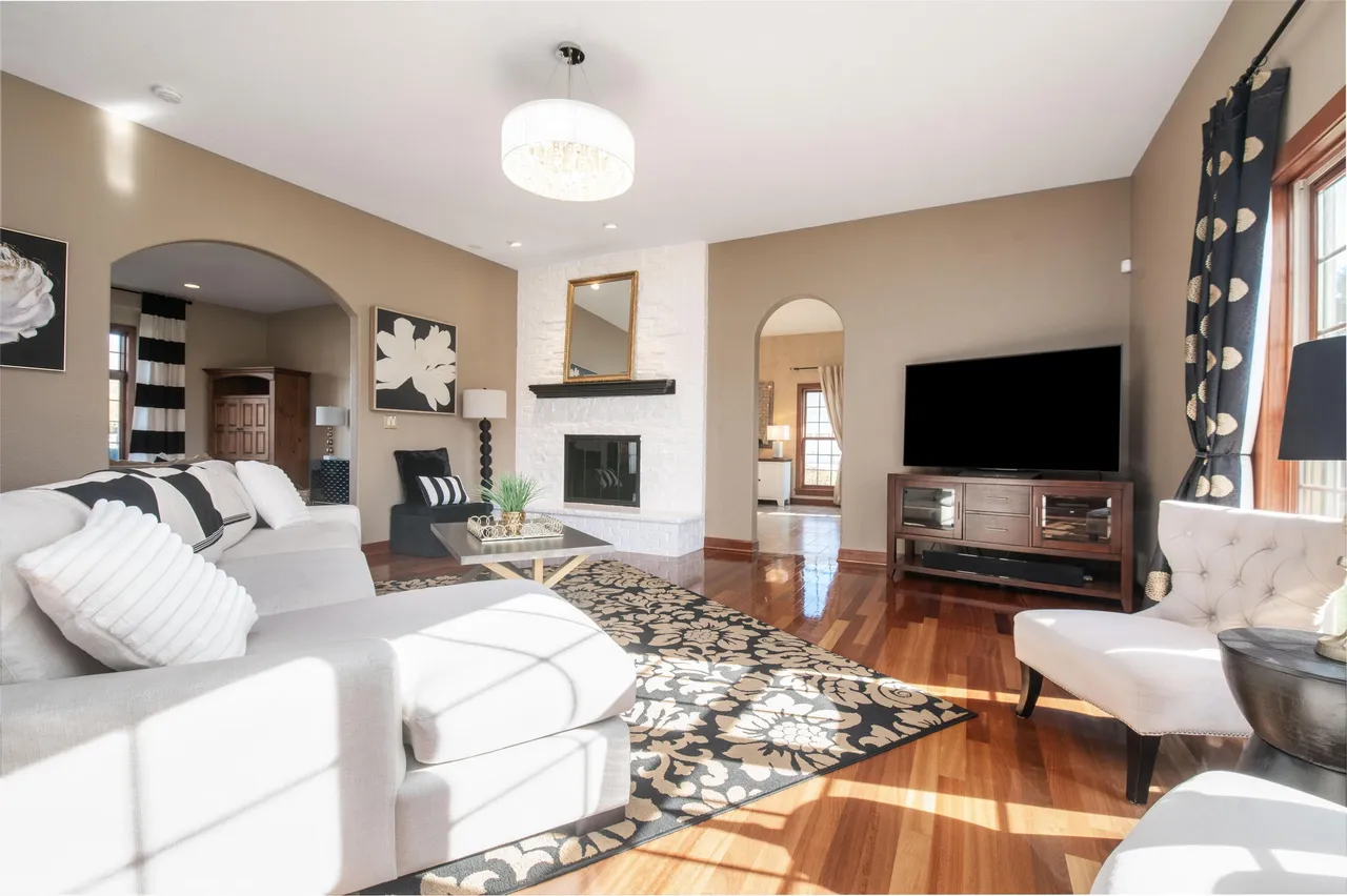 This is a warmly styled living room featuring a neutral color palette, hardwood floors, and a patterned area rug. The room is anchored by a white-painted brick fireplace with a decorative mirror above. A large television is situated on a wooden console table, and a comfortable sectional sofa and accent chairs provide ample seating.