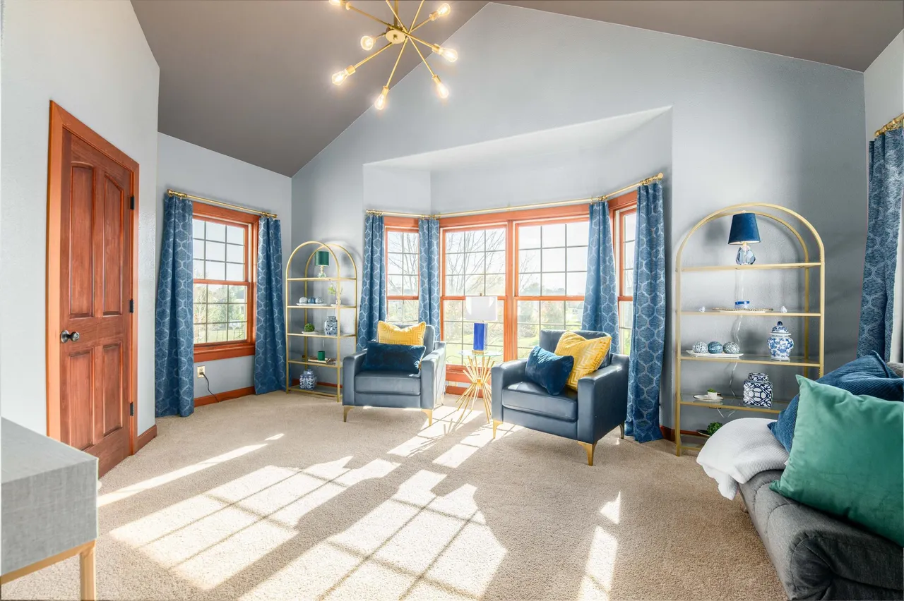 The living room features a cozy seating area with blue armchairs and decorative pillows, complemented by gold-framed bookshelves and window treatments. Sunlight streams through large windows, illuminating the neutral carpet and highlighting the room's architectural details, including a vaulted ceiling and contrasting ceiling paint. The overall impression is one of comfortable elegance and natural light.