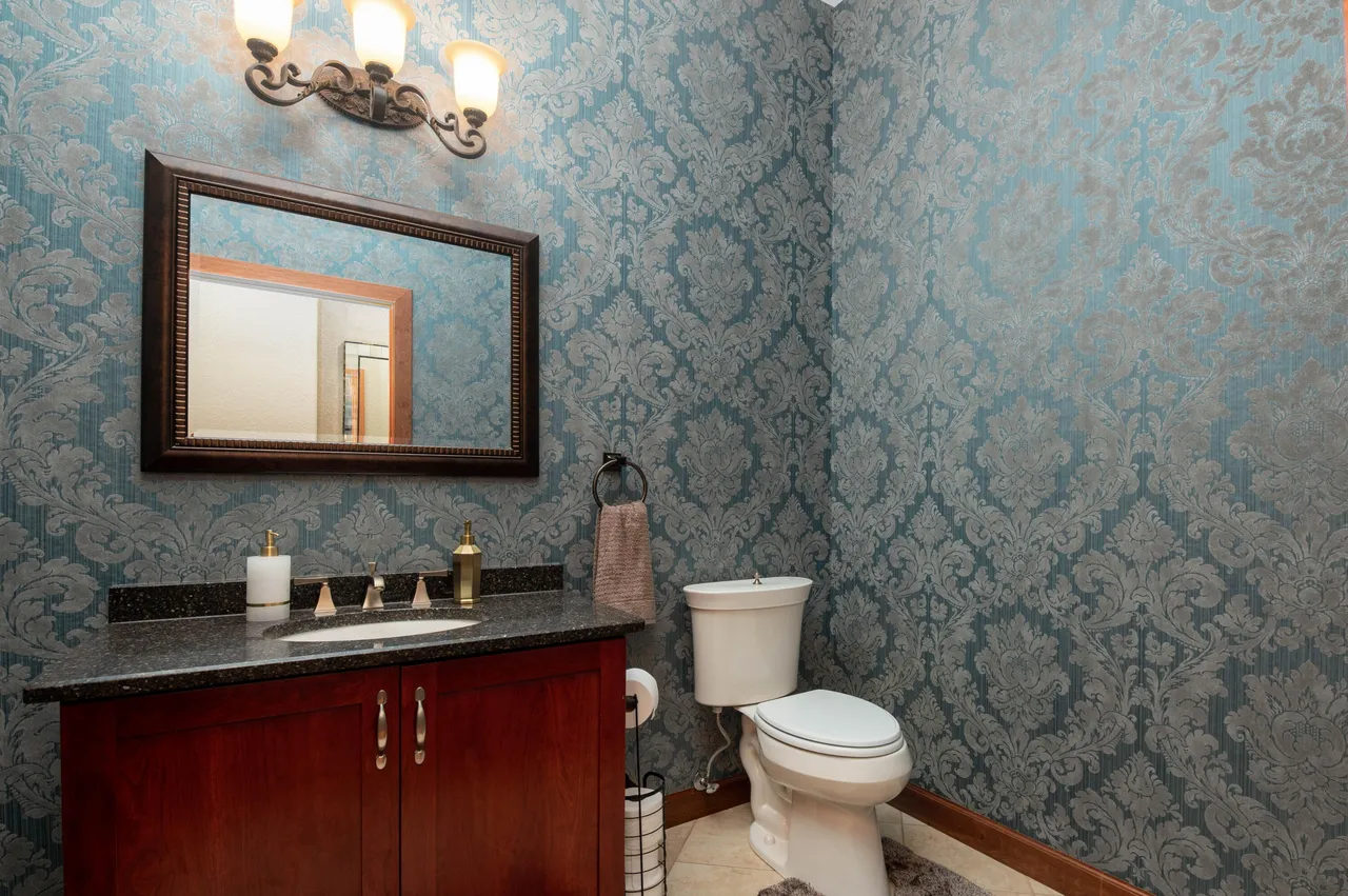 This is a guest bathroom with ornamental wallpaper and a cherry wood vanity with a dark countertop. A matching framed mirror is above the vanity, and the modern toilet sits next to a stand with toilet paper for easy access. A traditional light fixture is above the mirror, offering ample lighting.