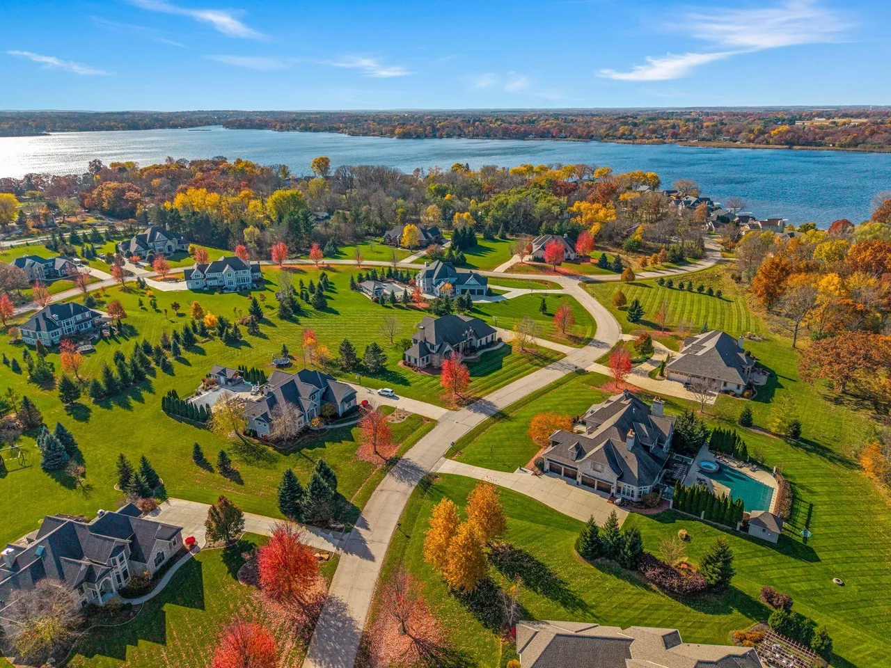 This aerial shot showcases a luxurious residential neighborhood with large, well-manicured lawns, mature trees displaying vibrant fall colors, and winding roads. Several impressive houses with varied architectural styles are visible, with some featuring swimming pools and spacious garages. In the background, a large blue lake adds to the idyllic setting, suggesting a desirable location with upscale living.