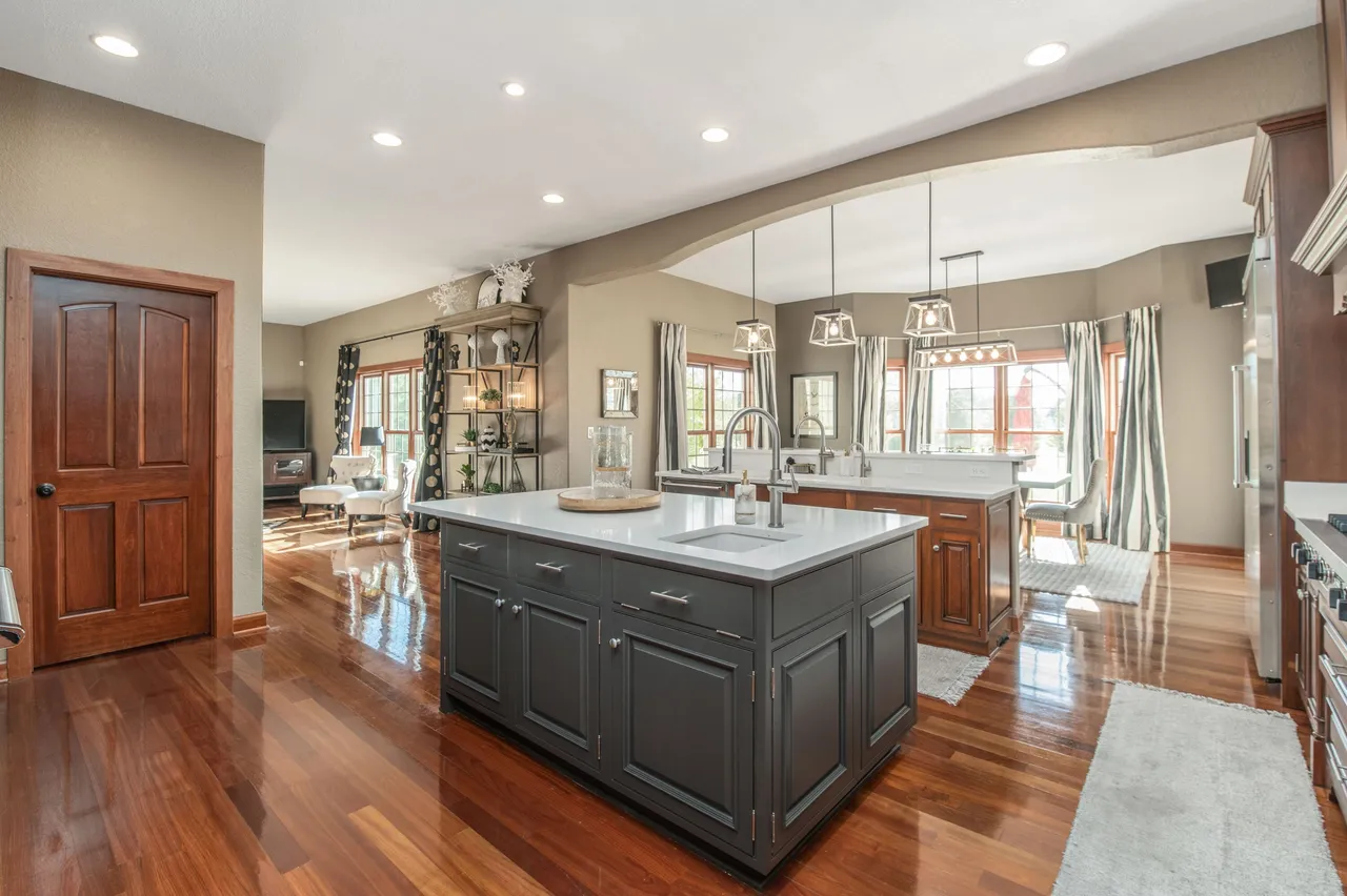This is a bright and spacious kitchen featuring hardwood floors and a large center island with a white countertop and gray cabinets. The kitchen also includes another island with darker wood cabinets and a breakfast nook area near large windows. Overall, the kitchen appears modern and well-maintained, offering ample space for cooking and entertaining.
