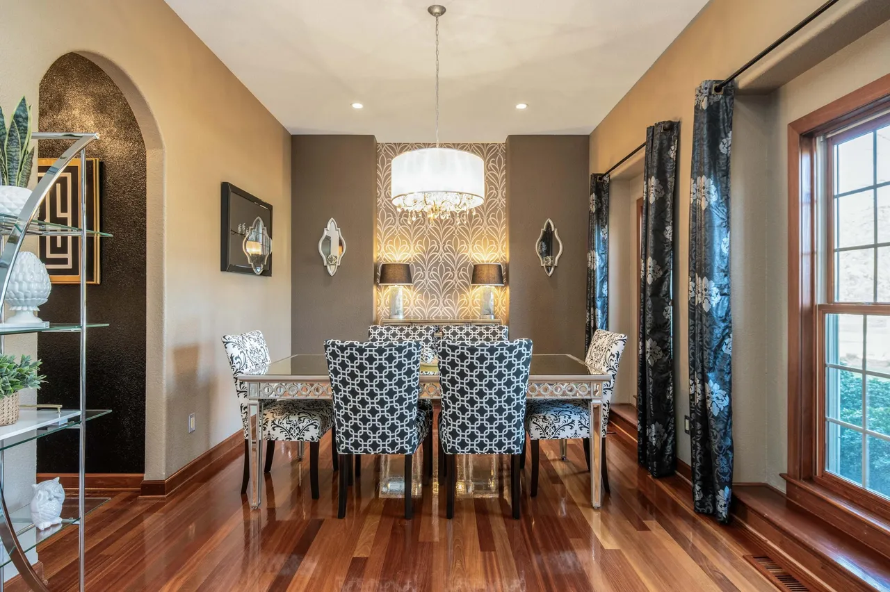 The image showcases a well-appointed dining room with a sophisticated ambiance. The focal point is a formal dining set with patterned upholstery, complemented by an elegant chandelier. The room features a decorative accent wall, rich hardwood flooring, and stylish window treatments, contributing to a luxurious and inviting atmosphere.