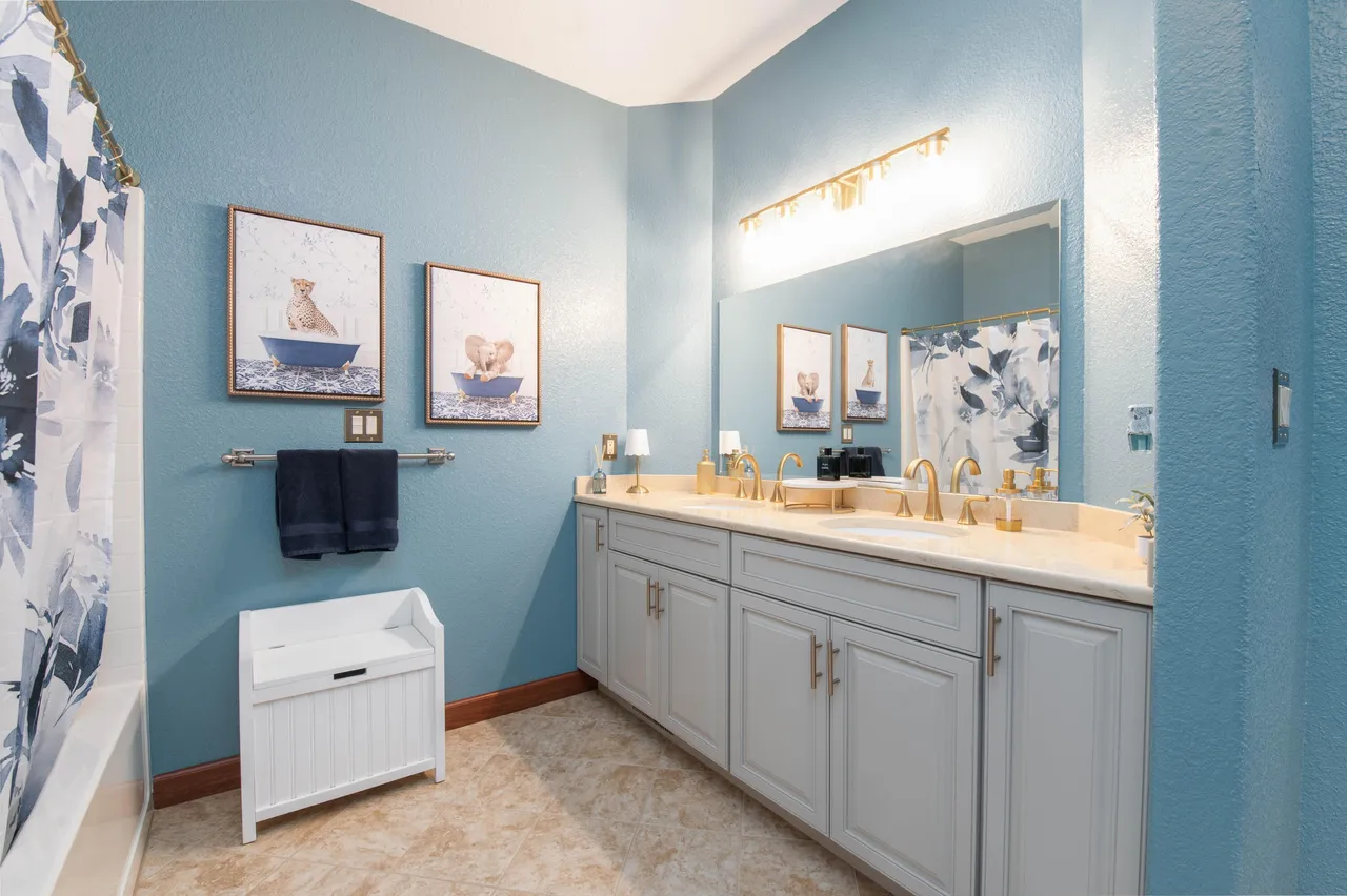 This is a well-lit guest bathroom featuring a double sink vanity with light gray cabinets and gold hardware. The walls are painted a calming blue, and decorative animal prints hang above a towel rack and bench. The room includes a shower-tub combination with a floral shower curtain and beige tile flooring.