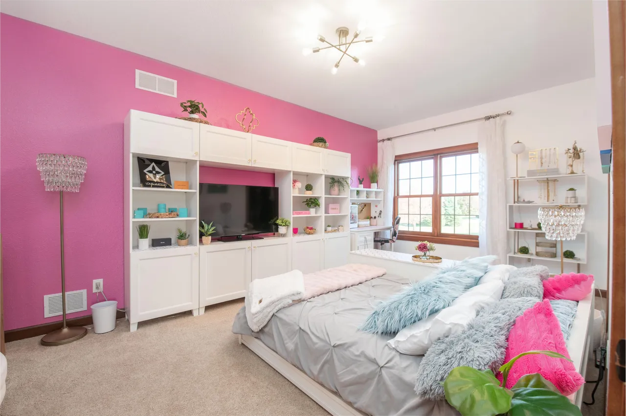This is a bright and stylish bedroom featuring a striking pink accent wall and built-in white shelving with a television. The room is furnished with a bed adorned with decorative pillows and throws, creating a cozy and inviting atmosphere. The decor elements and lighting fixtures add to the room's unique character.