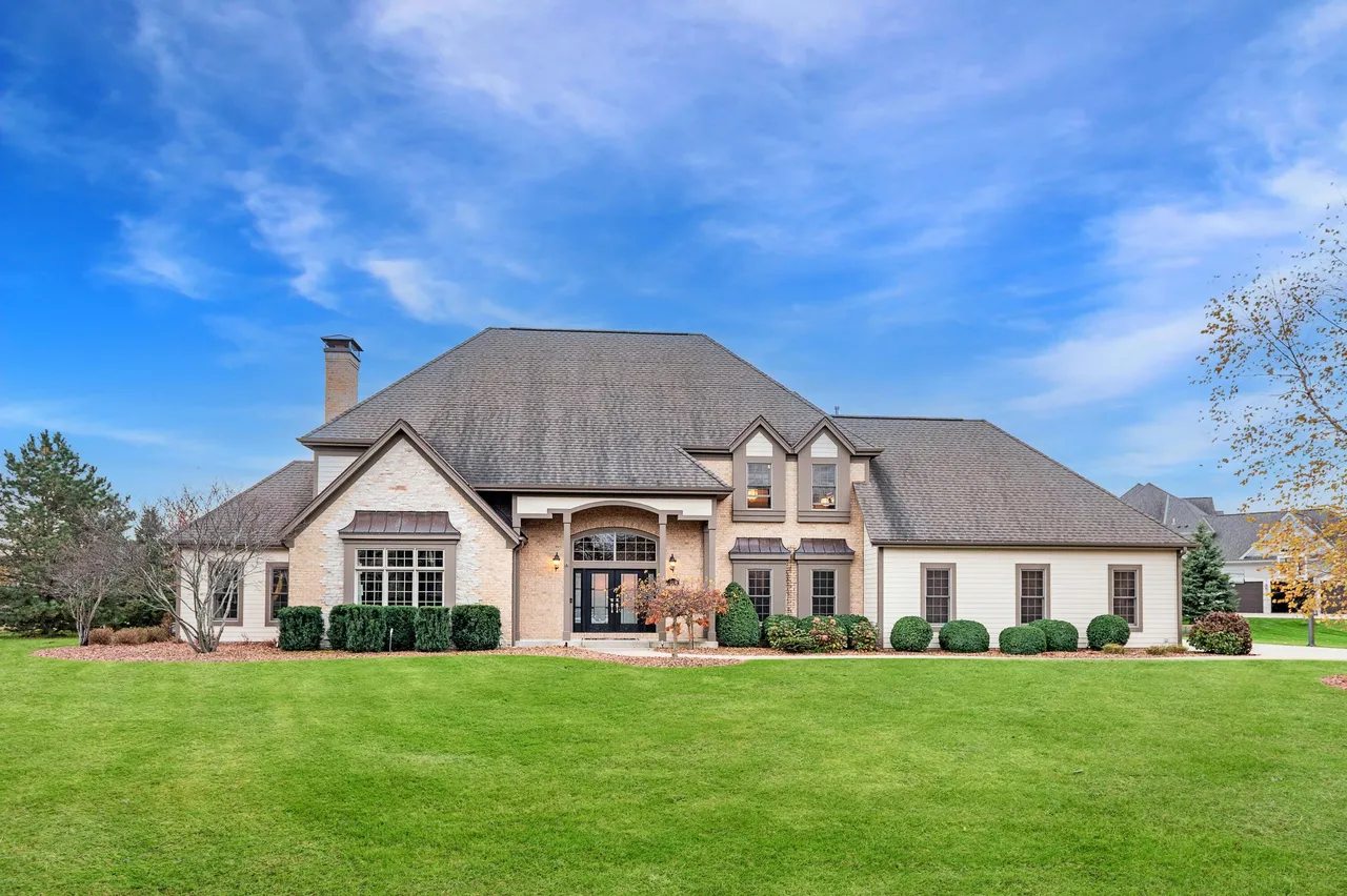 This is a front exterior shot of a large, luxurious home. The house features a brick and siding facade, a prominent roofline, and manicured landscaping. The expansive green lawn and well-maintained shrubs add to the property's curb appeal.
