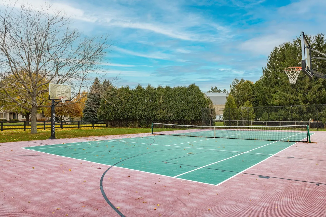 This image depicts a multi-purpose outdoor court, suitable for tennis and basketball. The court features a green playing surface with white lines and basketball hoops at either end. The surrounding area includes mature trees and greenery, enhancing the property's recreational appeal.