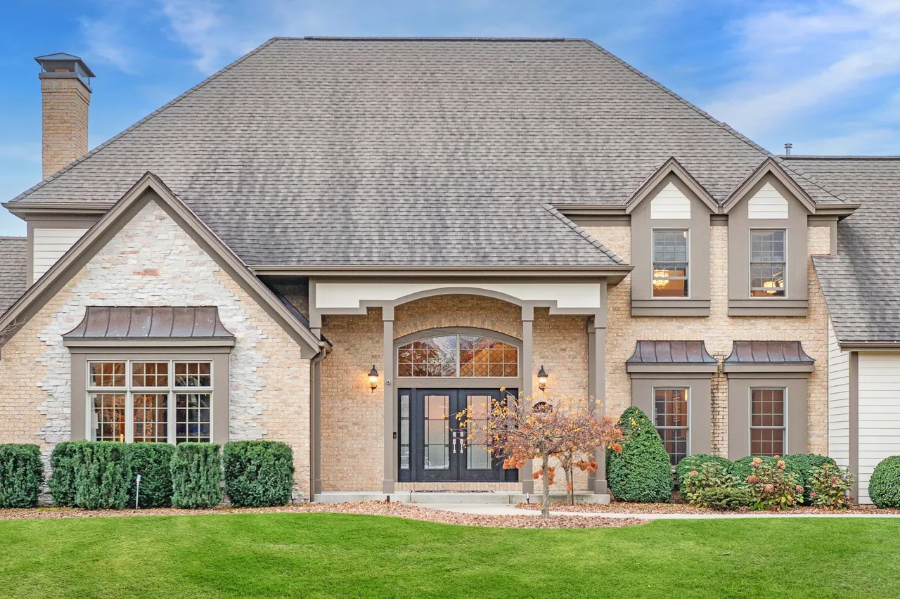 This is a front view of a well-maintained single-family home. The facade features a mix of brick and stone, a prominent asymmetrical gabled roof, and a symmetrical entrance. The lush green lawn and manicured bushes add to the property's curb appeal.