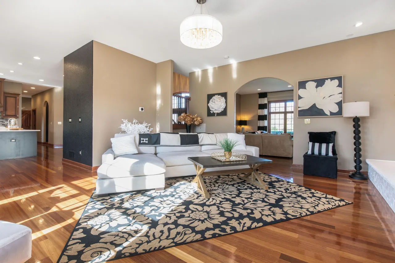The living room features hardwood floors, a white sectional sofa with decorative pillows, and a black and tan patterned area rug. A unique coffee table with black and gold accents sits in the center of the room. The walls are painted in a neutral beige tone, and two pieces of artwork adorn the walls.