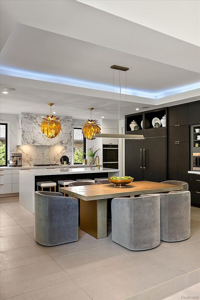 This is a modern kitchen with a large island, marble backsplash, and custom lighting fixtures. The kitchen features dark cabinetry on one side and white cabinetry on the other, creating a balanced contrast. A wooden dining table with gray upholstered chairs sits adjacent to the island, creating an inviting space for dining.