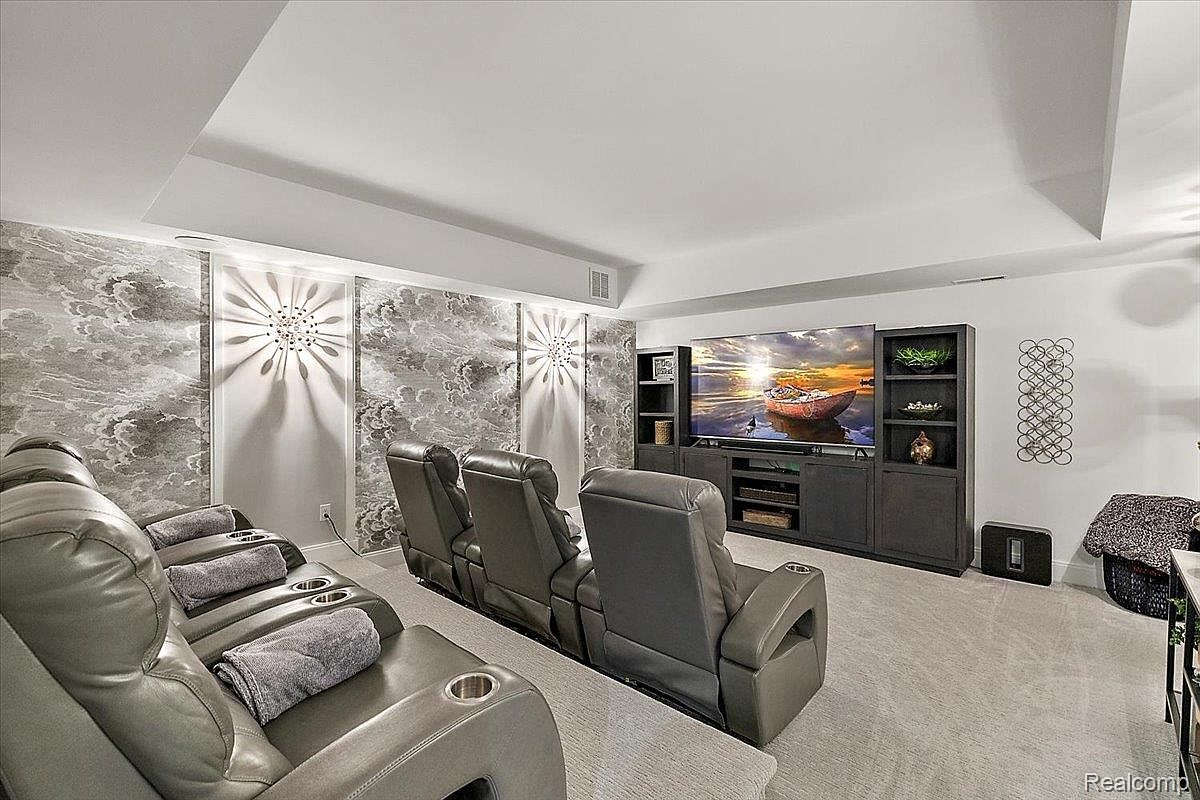 This is an interior shot of a home theater room. The room features tiered seating with leather recliners, a large flat-screen TV mounted on a dark entertainment center, and decorative wall panels with integrated lighting. The overall impression is one of comfort and modern luxury.