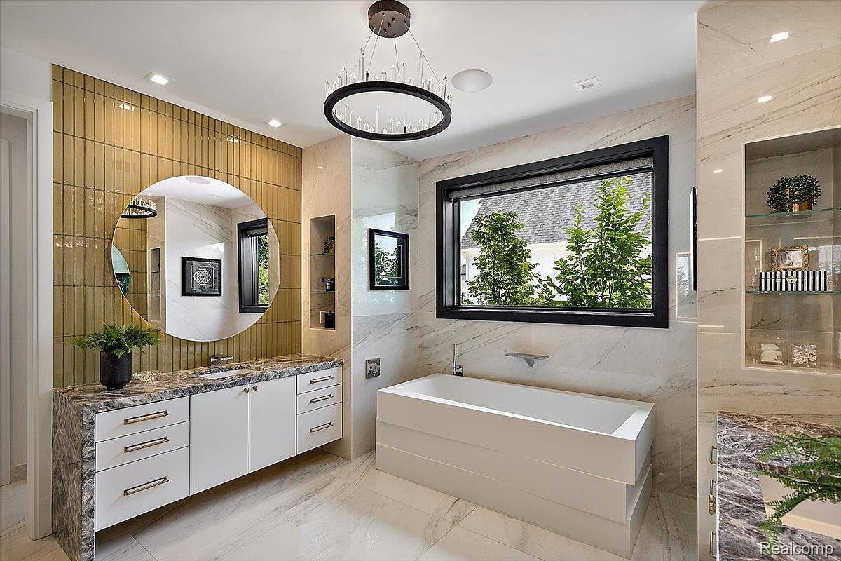 This is a luxurious primary bathroom featuring a modern freestanding bathtub positioned beneath a large window, offering natural light and a view of the outdoors. The vanity area showcases a marble countertop, white cabinetry with gold hardware, and a round mirror against a textured gold tile wall. A stylish chandelier hangs above, adding an elegant touch to the space.
