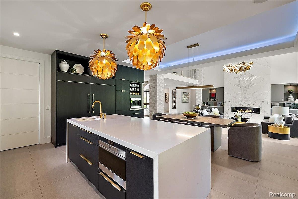This is a modern kitchen featuring a large island with a white countertop and dark cabinetry. The kitchen includes high-end appliances and unique pendant lighting. The space opens to a living area with a fireplace, creating an open and inviting atmosphere.