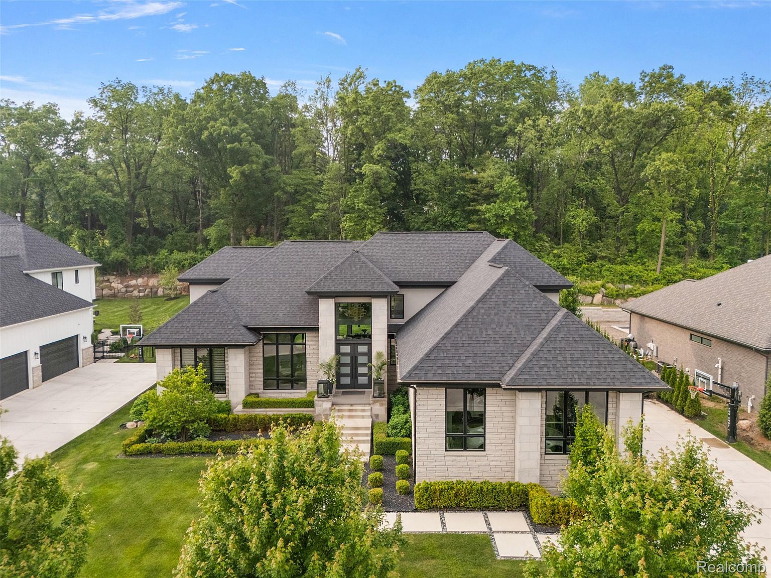 This aerial view showcases a luxurious, modern home with a dark gray roof, light stone facade, and black-framed windows. The property is surrounded by lush greenery and mature trees, providing privacy and a serene setting. The meticulously landscaped yard features manicured hedges, decorative stone pathways, and a well-maintained lawn, enhancing the home's curb appeal.