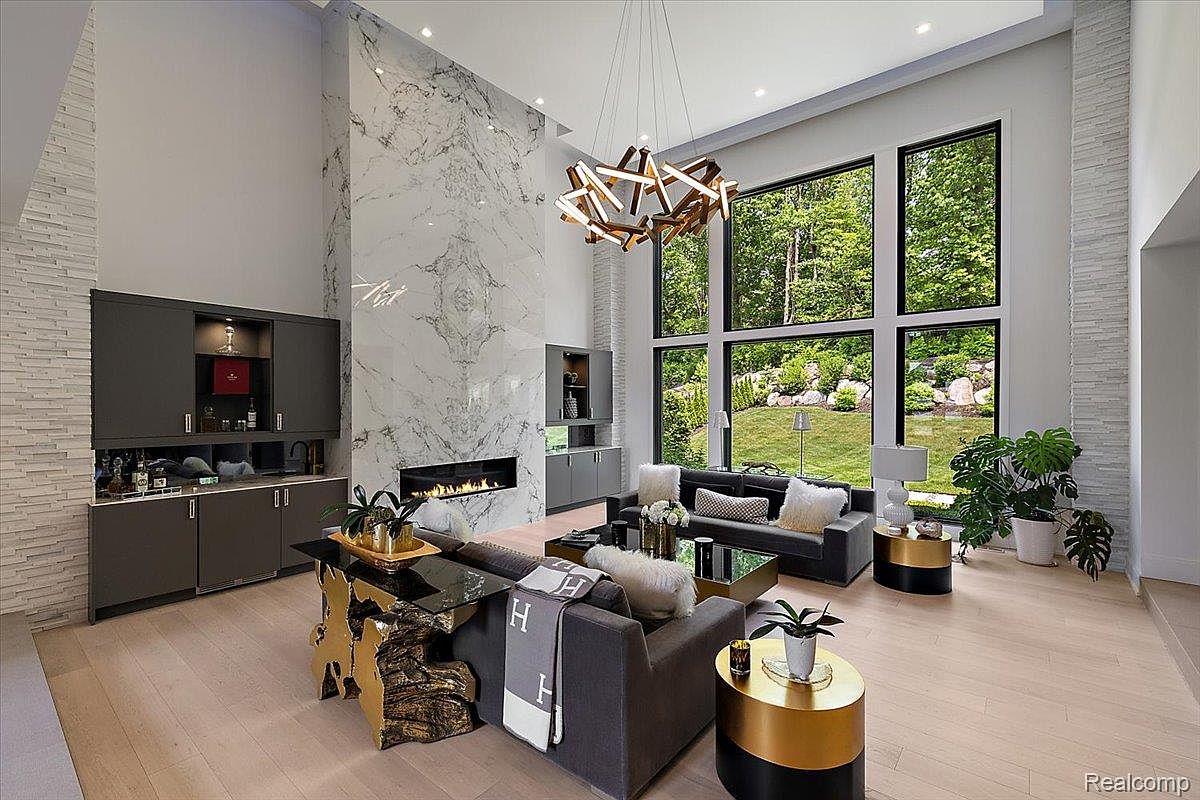 This is a spacious and modern living room featuring a high ceiling, a marble fireplace wall, and large windows offering a view of the lush greenery outside. The room is furnished with contemporary sofas, unique side tables, and a stylish chandelier, creating an elegant and inviting atmosphere. The light wood flooring complements the neutral color palette, enhancing the room's bright and airy feel.