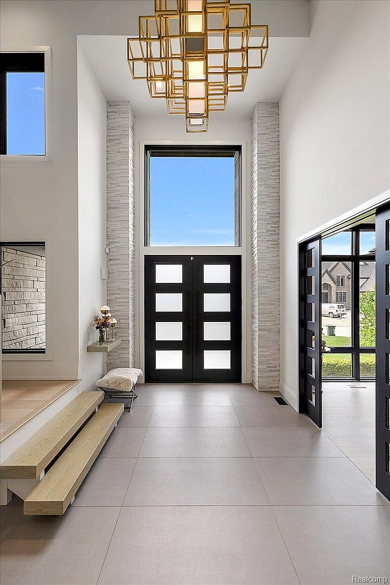 This is an interior shot of a modern home's entryway, featuring a high ceiling, a contemporary chandelier, and a large front door with sidelights. The flooring is light-colored tile, and there are minimalist wooden steps leading to a slightly raised area. The overall impression is one of spaciousness and modern elegance.