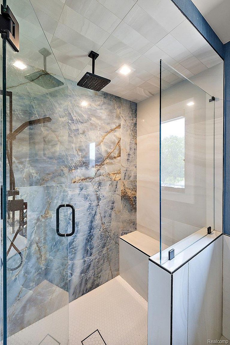 This is a modern primary bathroom featuring a walk-in shower with blue marble-patterned tile walls and a glass enclosure. The shower includes a rainfall showerhead and a handheld shower fixture. A built-in bench provides seating, and the floor is covered in small hexagonal tiles. The overall design is luxurious and spa-like.