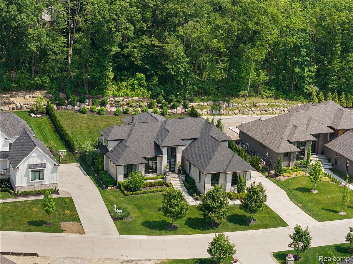 This aerial shot showcases a modern luxury home with a dark gray roof, light stone exterior, and meticulously landscaped grounds. The property features a long driveway, mature trees, and well-maintained lawns, creating a sense of privacy and elegance. The surrounding neighborhood is upscale, with similar high-end homes visible in the background.