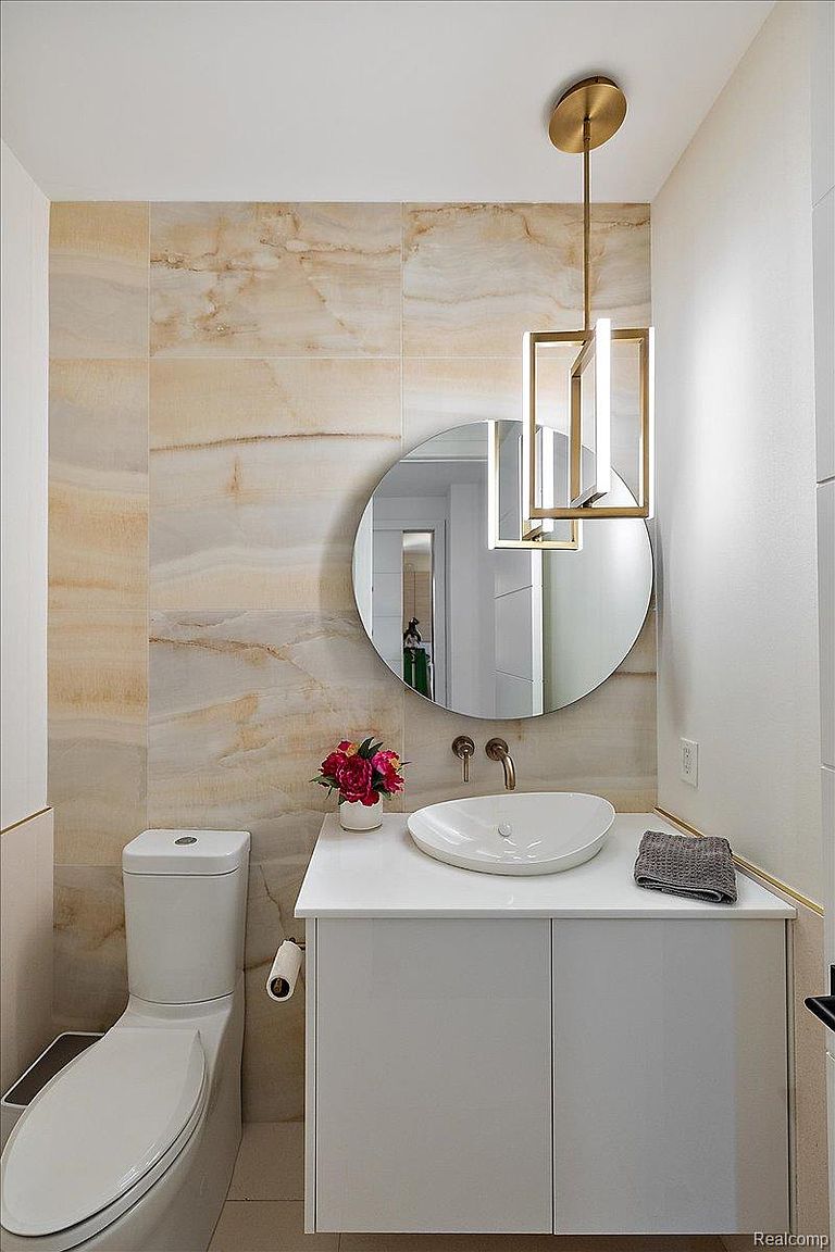 This is a well-lit, modern guest bathroom featuring a round mirror above a white vanity with a vessel sink. The walls are adorned with large, horizontally laid marble-patterned tiles, complemented by a contemporary gold-toned pendant light fixture. A toilet is visible to the left, and a gray towel adds a touch of contrast.