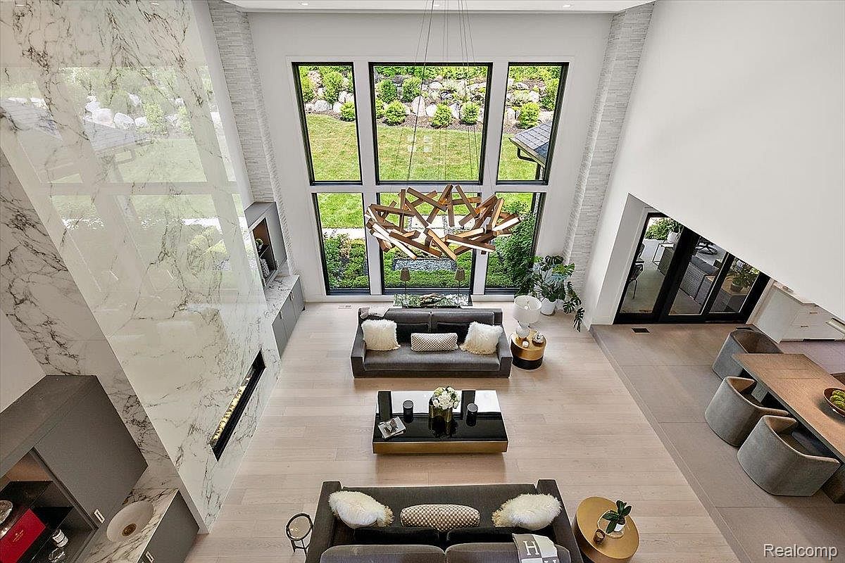 This is a high-angle shot of a modern living room with a soaring ceiling and expansive windows offering views of a lush garden. The room features two gray sofas, a black coffee table, and a striking geometric chandelier. A marble-clad fireplace wall adds a touch of luxury, while the light wood floors create a bright and airy atmosphere.