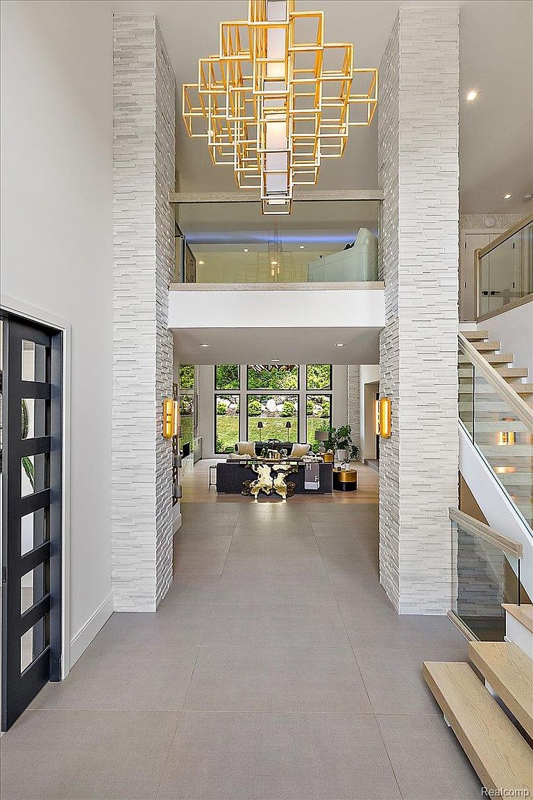 This interior shot showcases a grand foyer with a double-height ceiling and striking architectural details. The entrance features a modern chandelier, stone-clad pillars, and a view into the living room with large windows overlooking the backyard. A staircase with glass railings adds to the contemporary design, creating an open and inviting space.