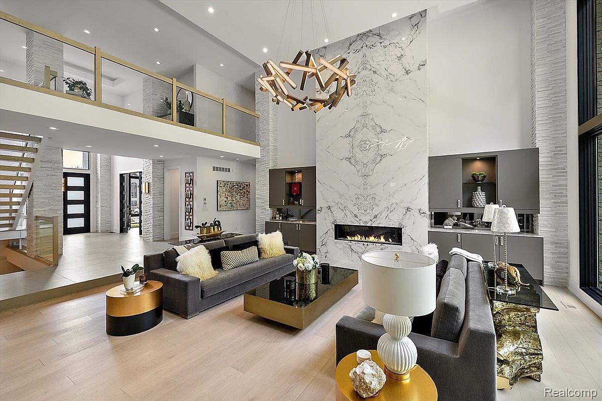 This is a spacious, modern living room featuring a soaring ceiling, a striking marble fireplace, and contemporary furnishings. The room is bathed in natural light from large windows, and a unique chandelier adds a touch of elegance. The open floor plan and neutral color palette create a sophisticated and inviting atmosphere.