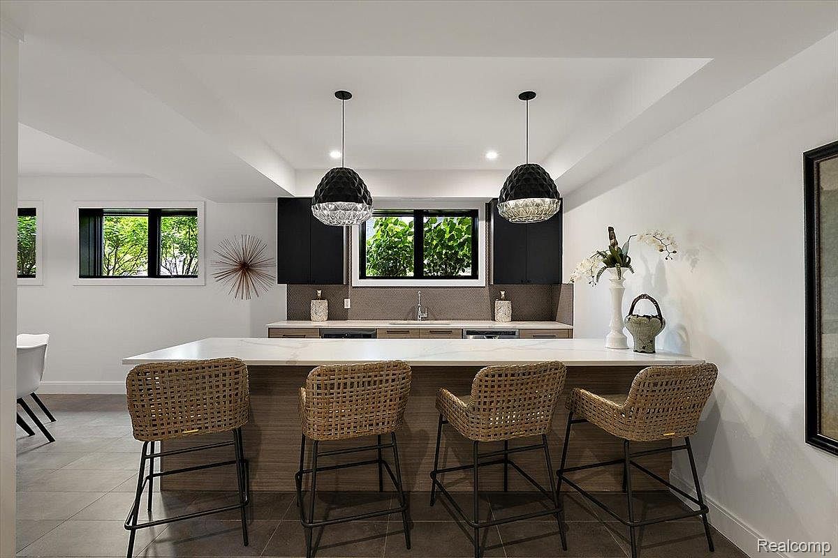 This is a modern kitchen featuring a large island with seating for four, illuminated by two pendant lights. The kitchen includes dark cabinetry and a window above the sink, providing natural light. The overall design is sleek and contemporary, creating an inviting space.