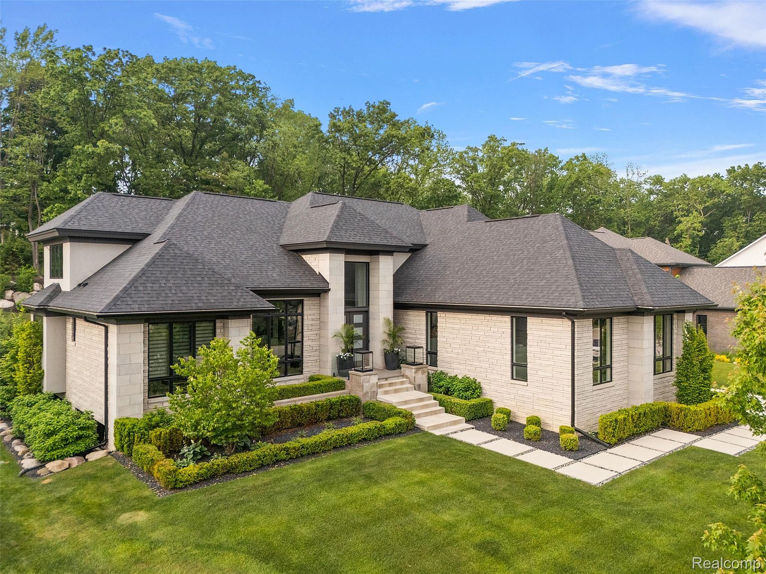 This is a front exterior view of a luxurious single-family home. The house features a light-colored brick facade, a dark gray roof, and black-framed windows. The manicured lawn and trimmed hedges add to the property's curb appeal, creating an inviting and sophisticated impression.
