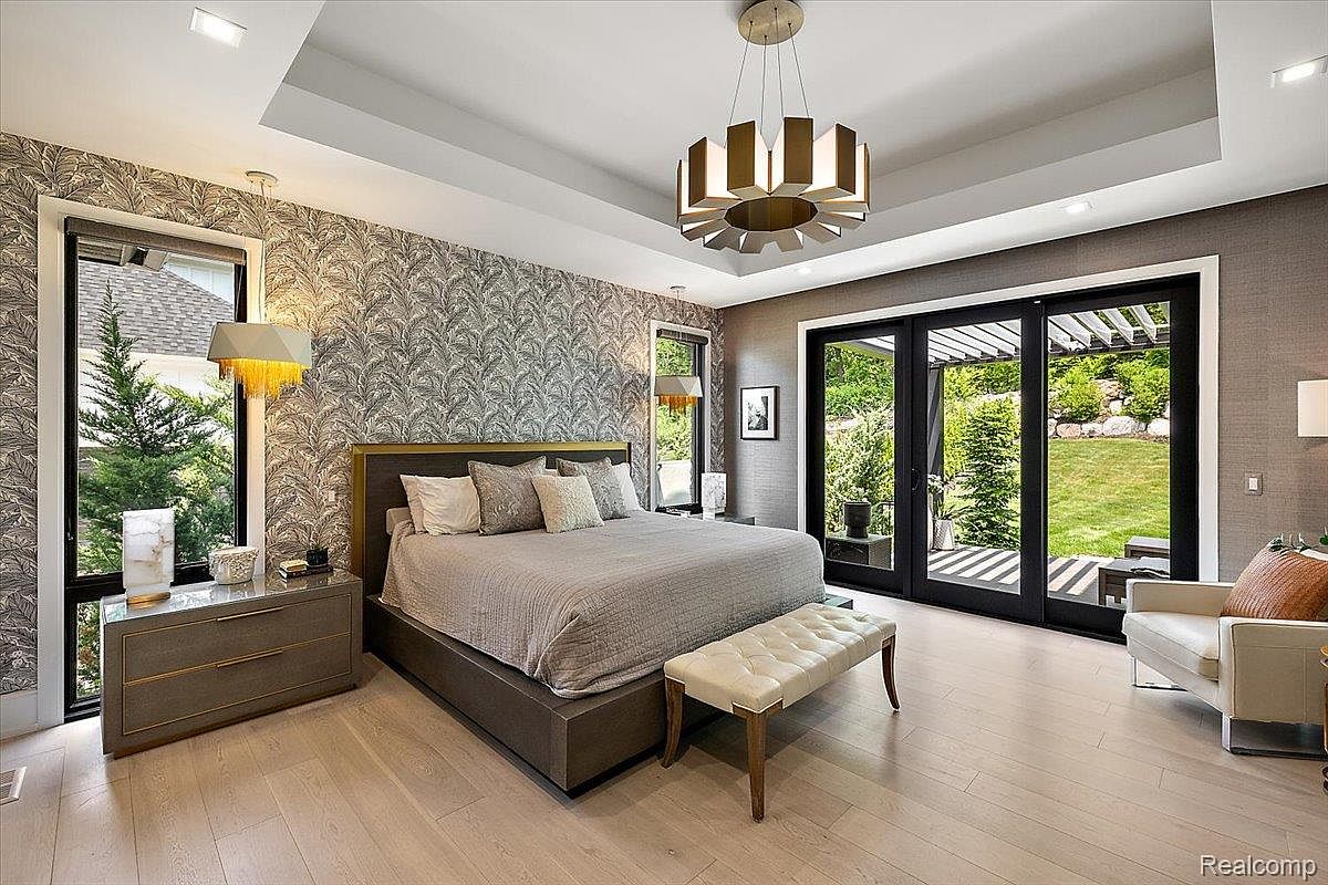 This is a primary bedroom featuring a king-size bed with a neutral-toned comforter and decorative pillows. The room has a modern aesthetic with patterned wallpaper on one wall and a large window offering natural light. A contemporary chandelier hangs from the tray ceiling, and sliding glass doors lead to an outdoor area, enhancing the room's spacious feel.