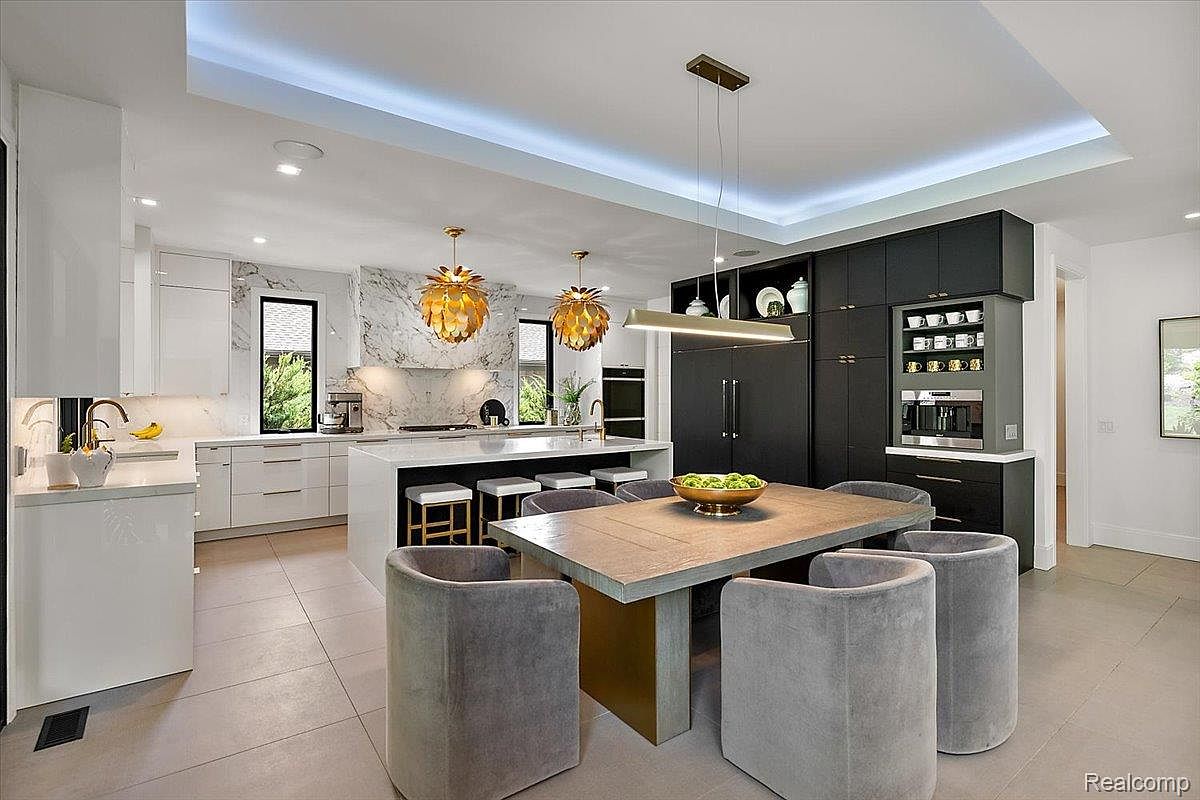 This is a modern kitchen with a large island, white cabinets, and marble backsplash. The kitchen also features black cabinets and stainless steel appliances. The dining area is adjacent to the kitchen, with a wooden table and gray chairs.