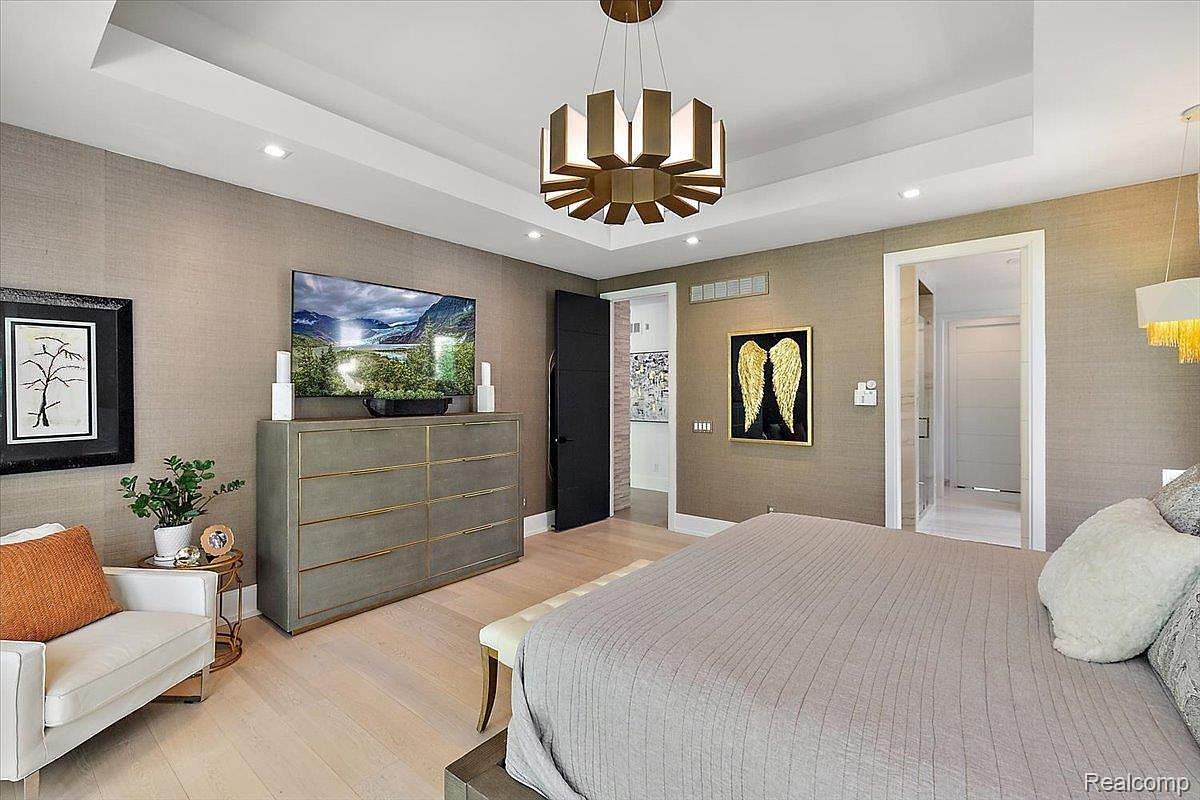 This is a primary bedroom featuring a king-size bed with a neutral-toned comforter, a gray dresser with a TV mounted above, and a modern chandelier. The walls are covered in a textured wallpaper, and there are two doorways, one leading to a bathroom and the other to a closet or hallway. The room has a sophisticated and calming atmosphere.