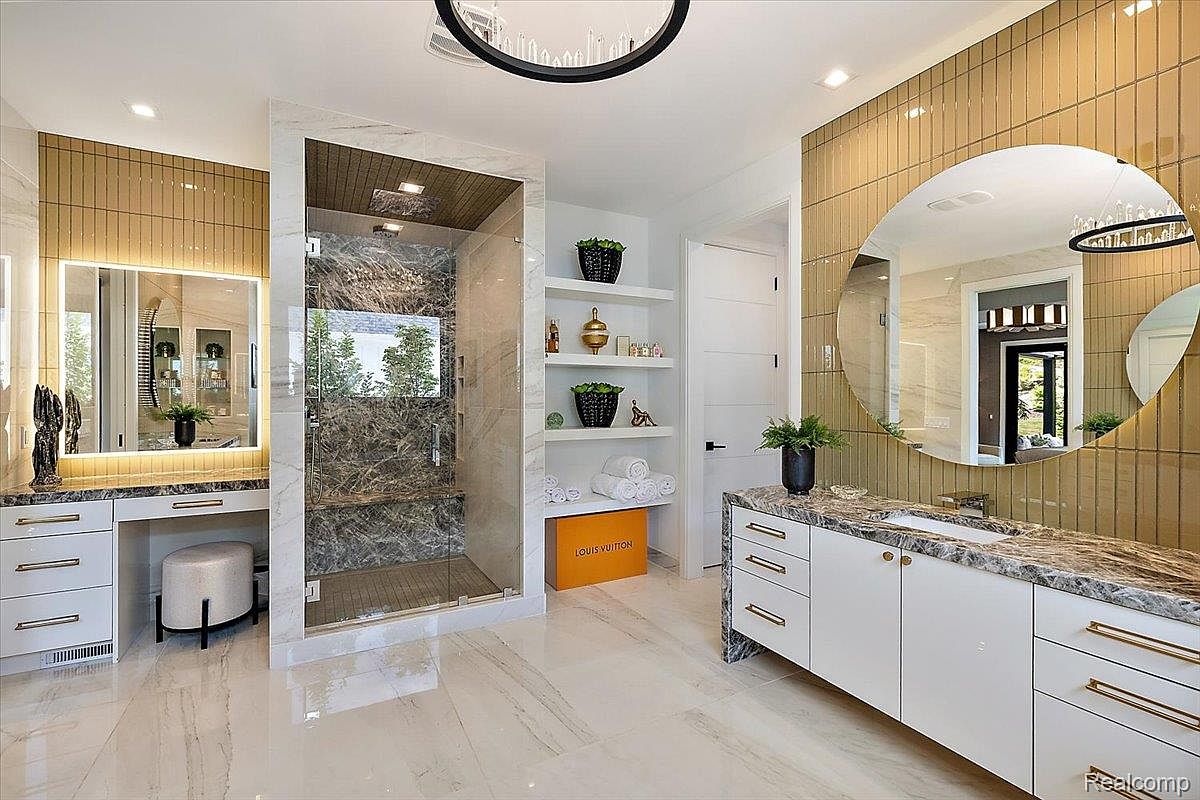 This is a luxurious primary bathroom featuring a walk-in shower with marble walls and glass doors, a vanity with a marble countertop and gold hardware, and a large round mirror. The walls are adorned with gold tiles, and the floor is a light-colored marble. The overall impression is one of elegance and sophistication.