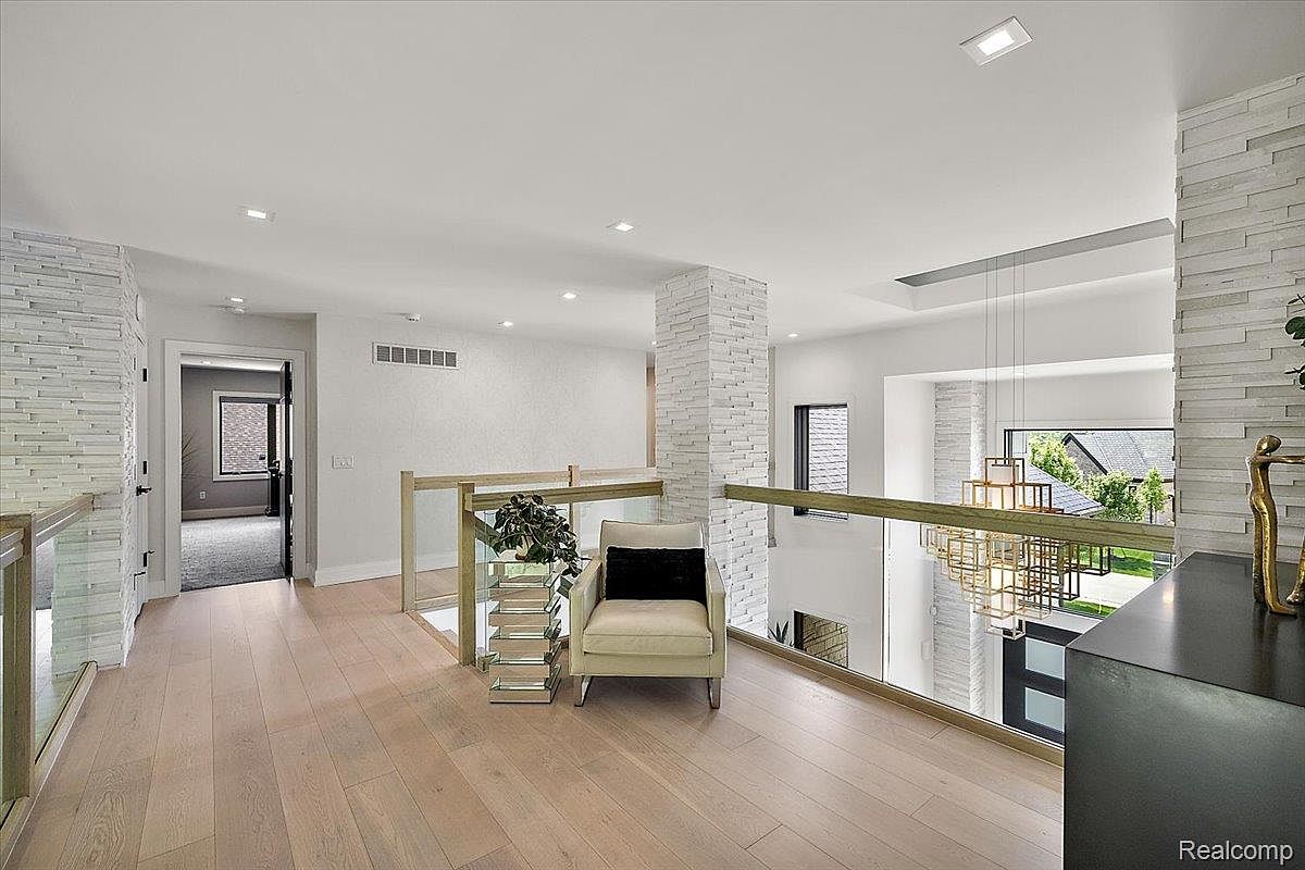 This is an interior shot of a modern hallway or landing area, featuring light wood flooring and a glass railing. A textured stone accent wall adds visual interest, and a contemporary light fixture hangs in the background. The space is well-lit and appears spacious, suggesting a high-end property.