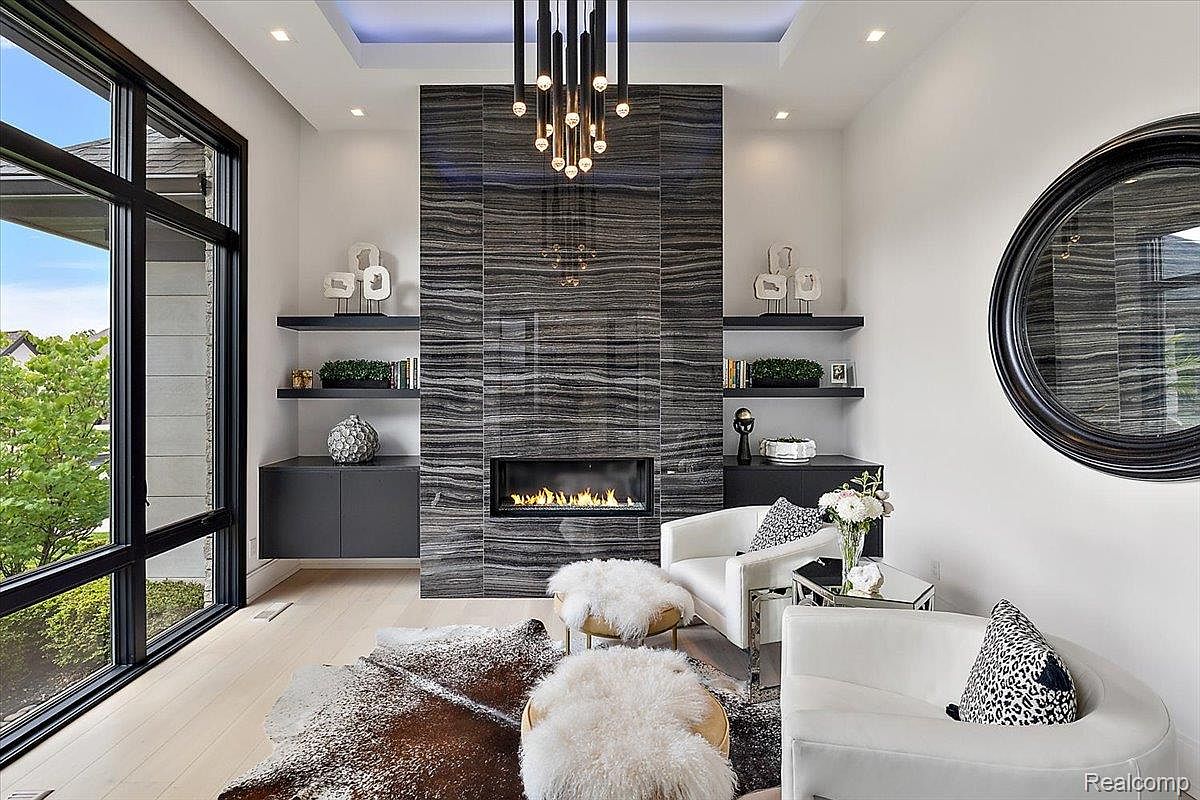 This is an interior shot of a modern living room featuring a striking fireplace with a dark, horizontally-striped tile surround. The room is furnished with white armchairs, decorative ottomans, and a cowhide rug, creating a luxurious and inviting space. The room also features built-in shelving and cabinets, a large window, and a unique chandelier.