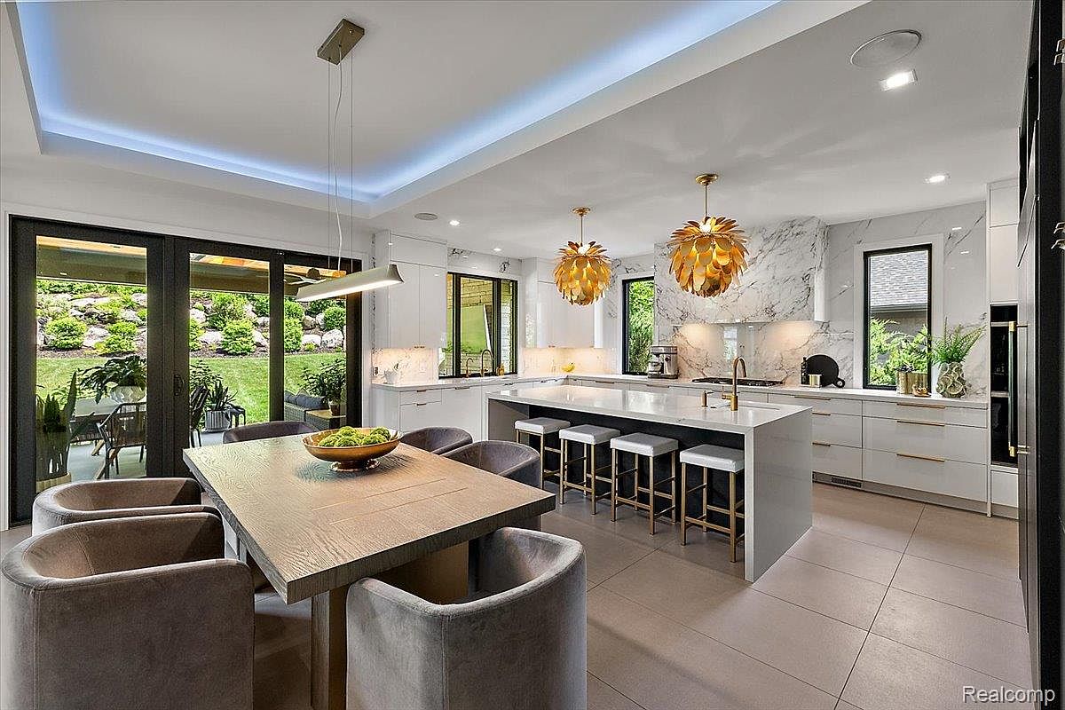 This is a bright and modern kitchen featuring white cabinetry, a large island with seating, and stylish gold pendant lighting. The kitchen also includes a dining area with a wooden table and gray upholstered chairs. Large sliding glass doors offer a view of the outdoor garden, creating a seamless indoor-outdoor living experience.