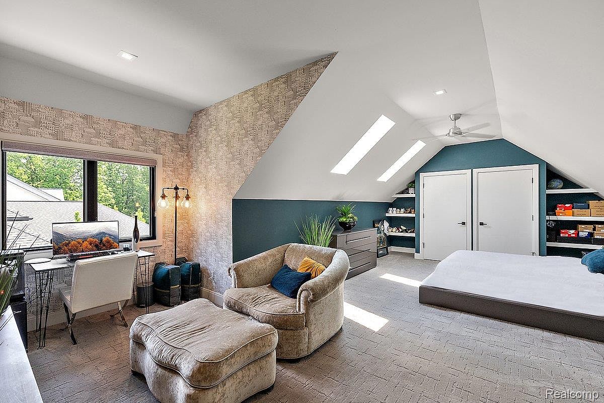This is a cozy attic bedroom featuring a unique architectural design with sloped ceilings and skylights. The room includes a work area with a desk, a comfortable armchair with an ottoman, and a bed. Built-in shelves and storage add functionality to the space, which is decorated in neutral tones with pops of color.