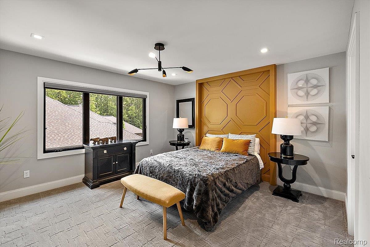 This is a well-staged bedroom featuring a queen-sized bed with a decorative headboard and a gray patterned comforter. The room is painted in neutral tones, complemented by a modern light fixture and matching bedside tables with lamps. A bench sits at the foot of the bed, and a window provides natural light, creating a cozy and inviting atmosphere.