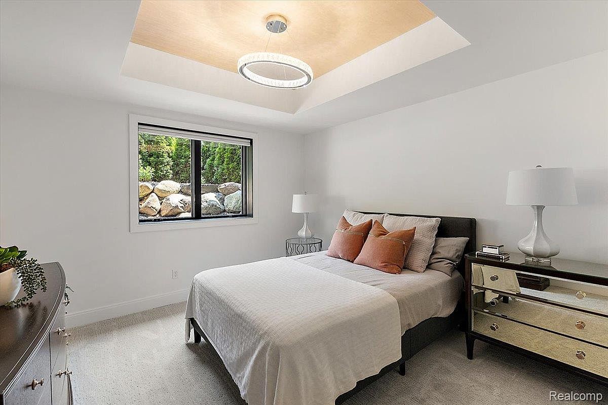 This is a well-lit bedroom featuring a bed with neutral bedding and orange accent pillows. A modern dresser with mirrored drawers sits to the right of the bed, and a window provides natural light and a view of greenery. The room has a clean and minimalist aesthetic, with white walls and a unique ceiling light fixture.