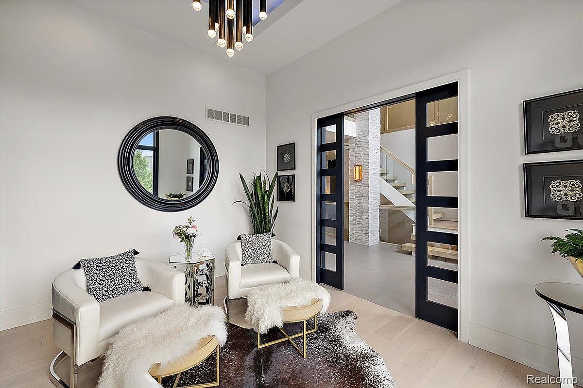 This is an interior shot of a modern living room featuring two white armchairs with decorative pillows, a round black-framed mirror on the wall, and a sliding black-framed glass door leading to a staircase. The room is decorated with a cowhide rug and small side tables, creating a stylish and inviting atmosphere. The perspective is from a medium distance, capturing the entire seating area and the doorway.