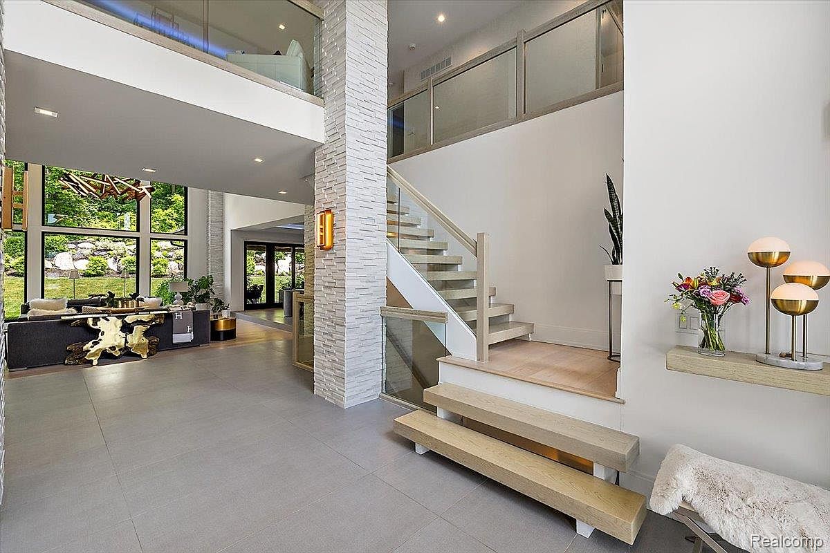 This interior shot showcases a grand hallway with a modern staircase. The hallway features light gray tile flooring, a striking stone-clad pillar, and a view into a living area with large windows overlooking a lush garden. The staircase has wooden steps and glass railings, leading to an upper level, complemented by contemporary lighting and decor.