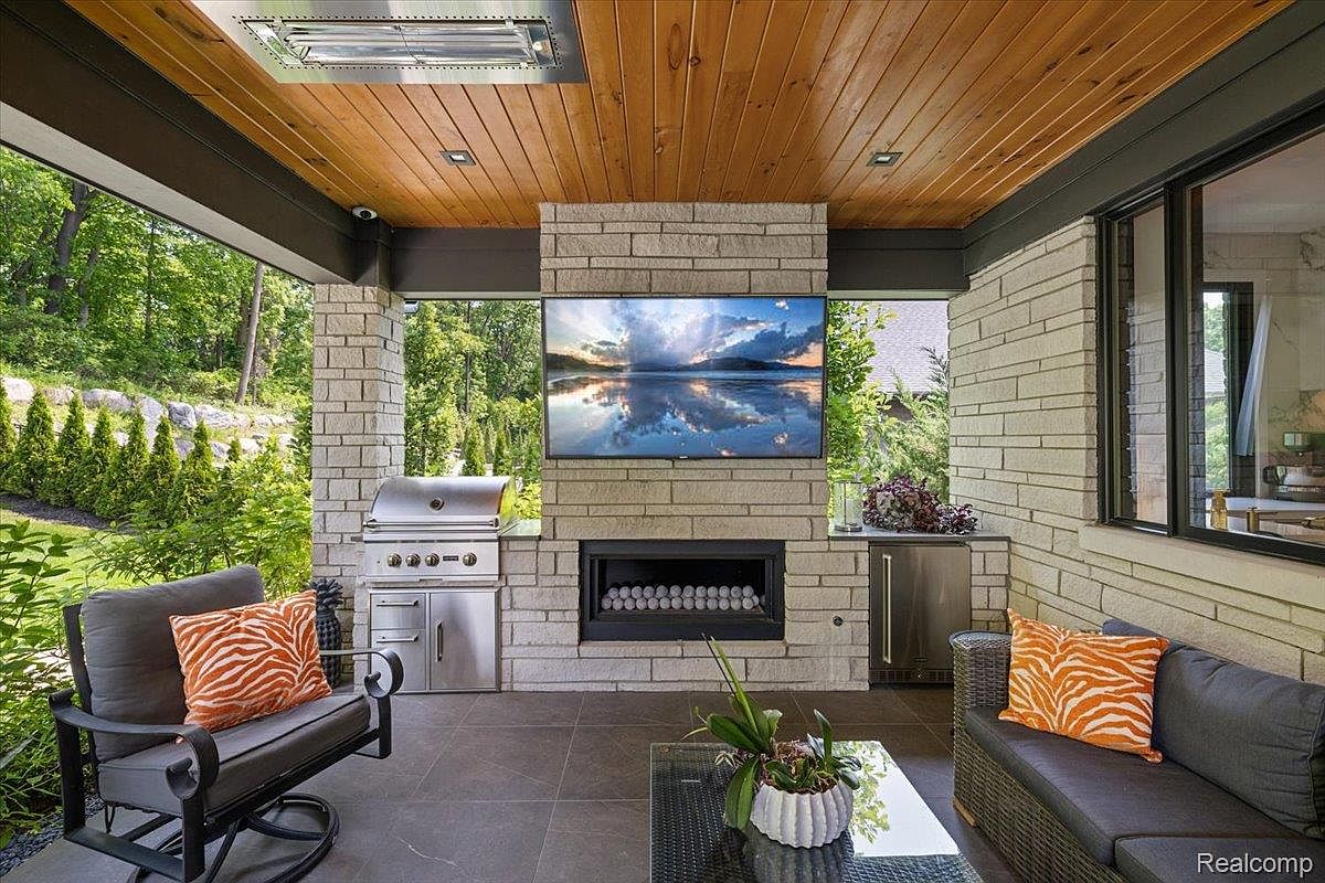 This image showcases a luxurious outdoor patio area, featuring a built-in grill, fireplace with a mounted television, and comfortable seating. The space is designed with stone accents and a wood-paneled ceiling, creating a sophisticated and inviting atmosphere. Lush greenery surrounds the patio, providing privacy and a serene backdrop.