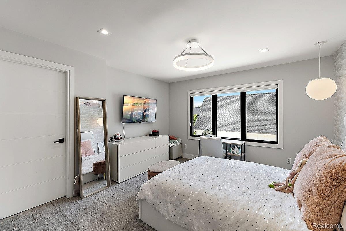 This is a bright and airy bedroom featuring a large window with black frames, a modern white dresser with a television mounted above, and a full-length mirror. The bed is neatly made with white linens and soft pink pillows, complemented by a round ottoman. The room is decorated in neutral tones, creating a calming and inviting atmosphere.
