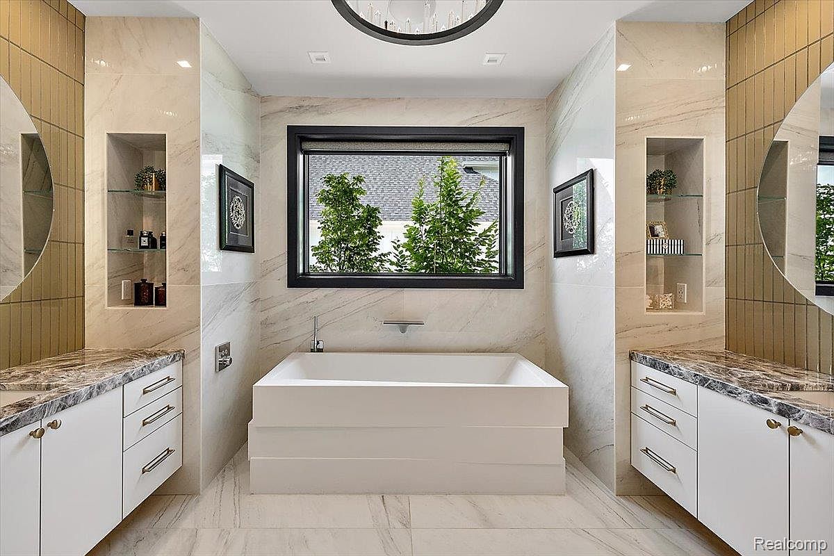 This is a luxurious primary bathroom featuring a modern freestanding bathtub centered under a window with a view of greenery. The walls are adorned with marble-like tiles, and there are built-in shelving units on either side of the room. The vanity areas have marble countertops and white cabinets, creating a clean and elegant aesthetic.