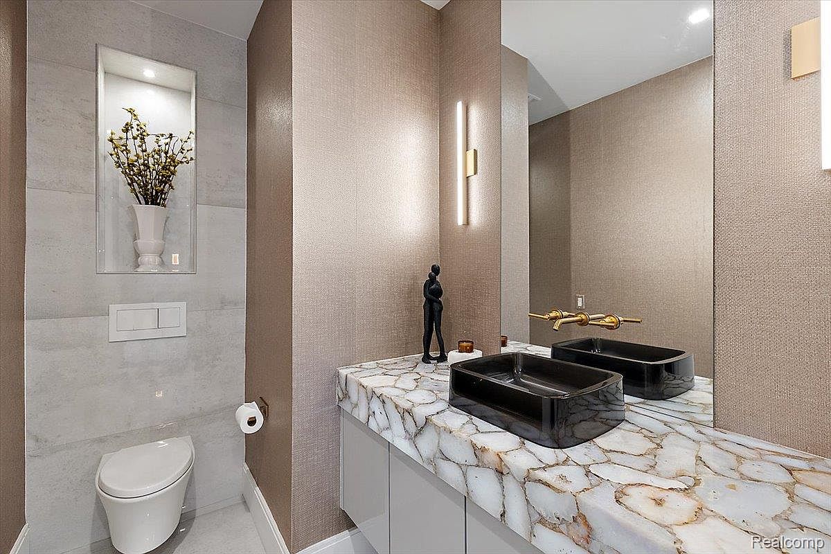 This is a modern guest bathroom featuring a unique quartz countertop with two black rectangular vessel sinks and gold-toned faucets. The walls are covered in a textured wallpaper, complemented by a vertical light fixture. A toilet is tucked into a corner with a decorative niche above, adding a touch of elegance to the space.