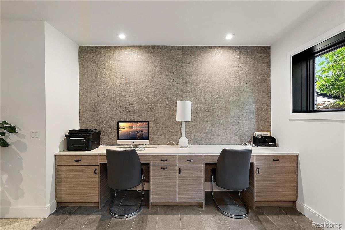 This is an interior shot of a home office featuring a built-in desk with cabinets and drawers, providing ample storage. The workspace is illuminated by recessed lighting and a table lamp, with a computer and printer set up for functionality. The room has a modern aesthetic with a textured accent wall and a window offering natural light.