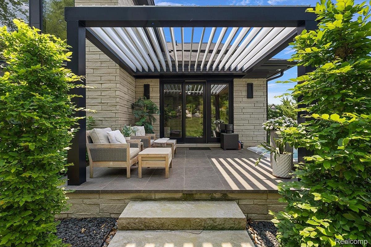 This is a well-designed outdoor patio area featuring a modern pergola with white slats and black supports, providing shade over a seating area. The patio is furnished with comfortable chairs and a small table, set on a stone-tiled surface. Lush greenery flanks the sides, adding to the privacy and aesthetic appeal of the space, while stone steps lead down to a gravel area.