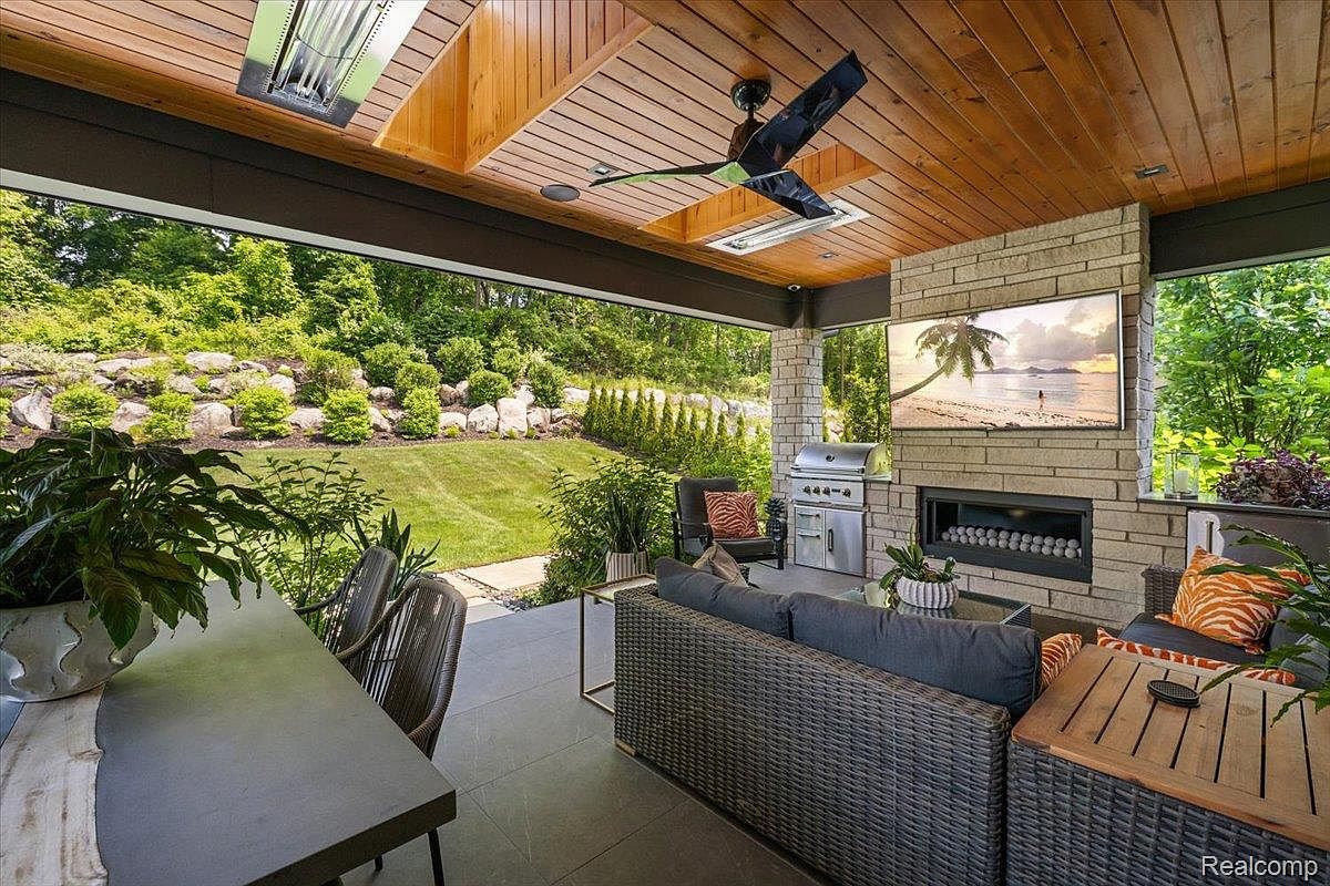 This image showcases a luxurious outdoor living space featuring a covered patio with a wood-paneled ceiling and a ceiling fan. A stone fireplace with a mounted television serves as a focal point, complemented by comfortable wicker seating and a built-in grill. The patio overlooks a well-manicured lawn and landscaped garden, creating an inviting and upscale outdoor retreat.