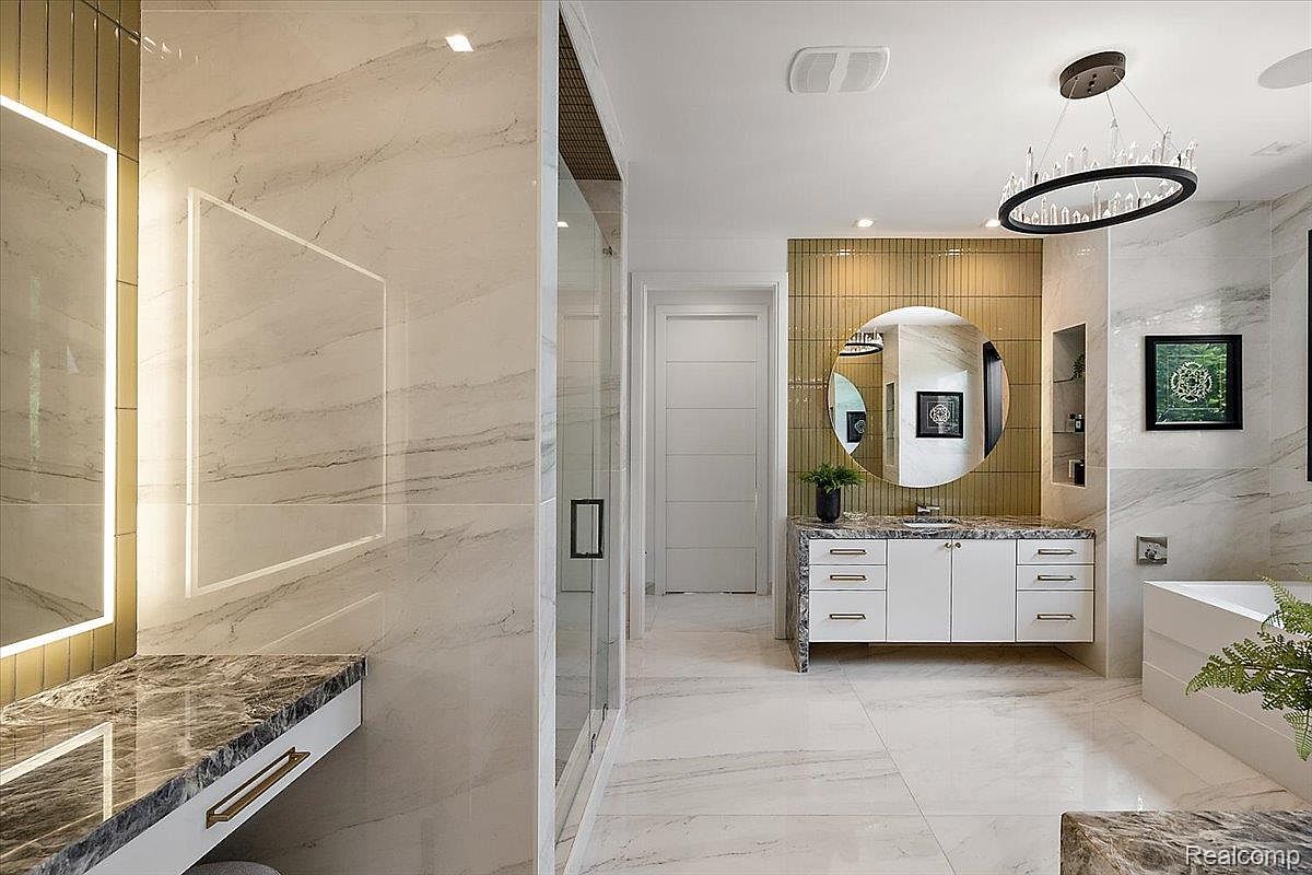 This is a luxurious primary bathroom featuring marble walls and flooring, a modern vanity with a marble countertop, and a large round mirror illuminated by a chandelier. A glass-enclosed shower is visible to the left, and the overall design exudes elegance and sophistication. The perspective is from the entrance of the bathroom, showcasing the layout and high-end finishes.