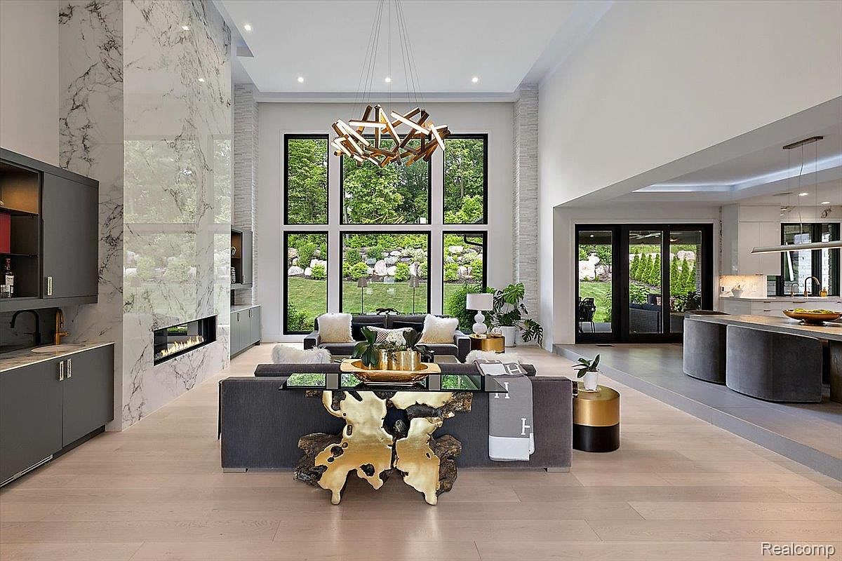 This is a spacious and modern living room featuring a double-height ceiling and a wall of windows overlooking a lush garden. The room is furnished with a gray sofa, armchairs, and a unique gold-colored accent table. A marble fireplace and contemporary chandelier add to the luxurious feel of the space, which seamlessly transitions into an open-concept kitchen area.